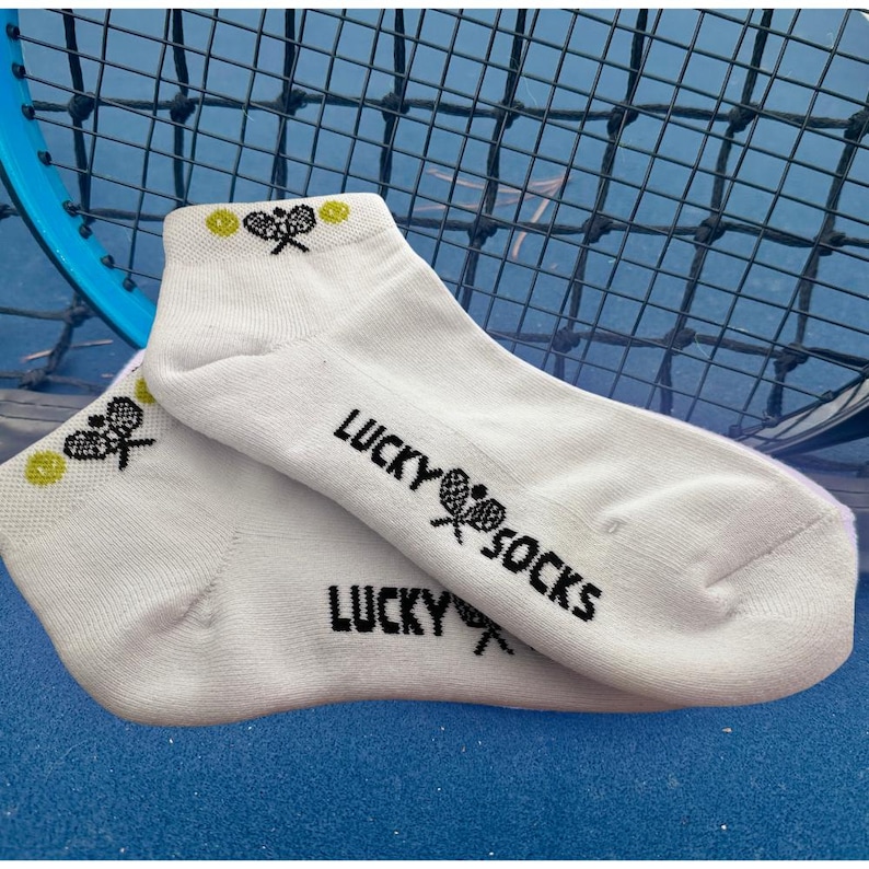 Puede incluir: Calcetines tobilleros blancos con el texto "LUCKY SOCKS" en negro. Los calcetines presentan un dise&ntilde;o de raqueta y pelota de tenis. Una raqueta y una red de tenis son visibles en el fondo.