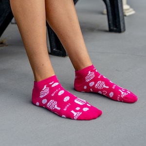 May include: Bright pink ankle socks with a white pattern of various food items and the word "binki" printed on the side. The socks are worn by a person sitting down.