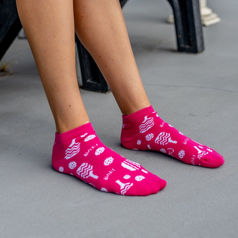 May include: Bright pink ankle socks with a white pattern of various food items and the word "binki" printed on the side. The socks are worn by a person sitting down.