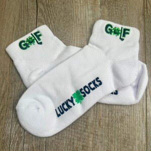 May include: White ankle socks with the word "GOLF" embroidered in green and a shamrock design on the cuff. The socks also feature the text "LUCKY SOCKS" in dark blue and green.
