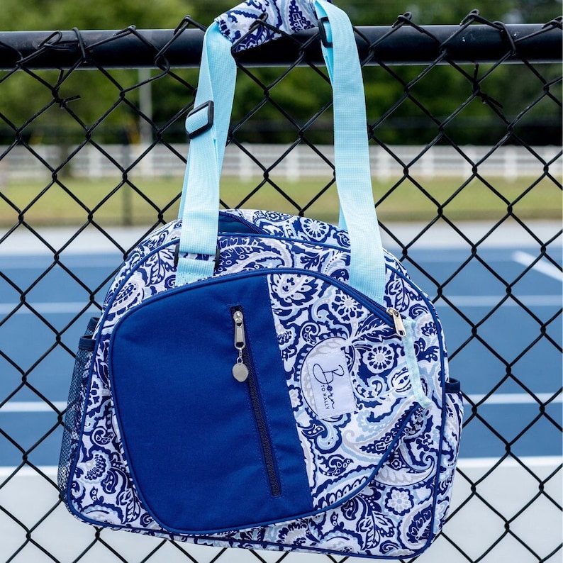 May include: A blue and white paisley patterned tennis bag with a zipped pocket and a blue strap. The bag has the text "Born to Rally" on it.