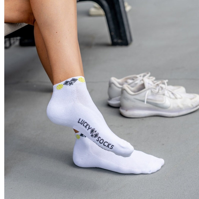 May include: White ankle socks with the text "LUCKY SOCKS" in black. The socks have a small design in black and yellow near the top. A pair of white sneakers is in the background.