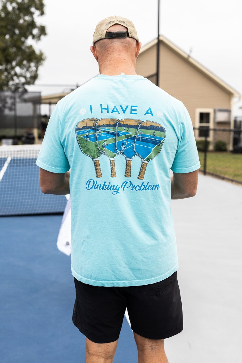 K&ouml;nnte beinhalten: Ein hellblaues T-Shirt mit einem Aufdruck von vier Pickleball-Schl&auml;gern und dem Text "Ich habe ein Dinking-Problem".