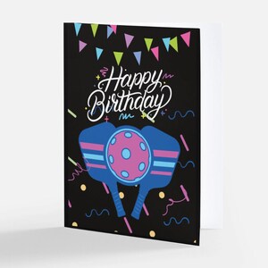 K&ouml;nnte beinhalten: Eine schwarze Karte mit einem farbenfrohen Design, das zwei blaue Pickleball-Schl&auml;ger mit einem rosa Ball in der Mitte zeigt. Der Text "Happy Birthday" ist in wei&szlig;er Kursivschrift auf der Karte geschrieben.