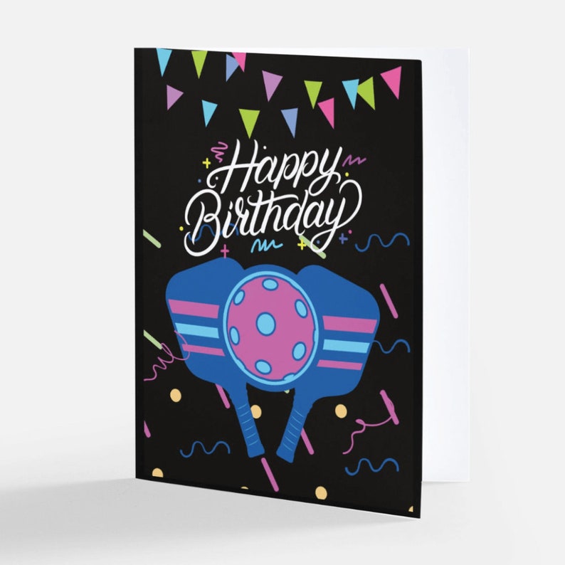 K&ouml;nnte beinhalten: Eine schwarze Karte mit einem farbenfrohen Design, das zwei blaue Pickleball-Schl&auml;ger mit einem rosa Ball in der Mitte zeigt. Der Text "Happy Birthday" ist in wei&szlig;er Kursivschrift auf der Karte geschrieben.
