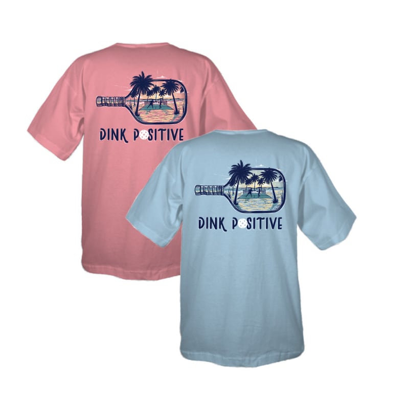 K&ouml;nnte beinhalten: Zwei T-Shirts mit einem Pickleball-Schl&auml;ger-Design. Der Schl&auml;ger befindet sich in einer Flasche mit einer Strandszene im Inneren. Der Text auf der R&uuml;ckseite der Shirts lautet "Dink Positive". Ein Shirt ist rosa und das andere hellblau.