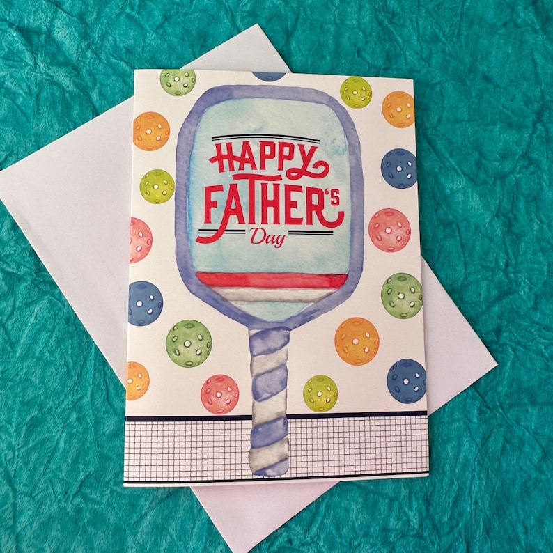 K&ouml;nnte beinhalten: Eine wei&szlig;e Karte mit einer Illustration eines blauen und wei&szlig;en Paddleball-Schl&auml;gers. Der Paddleball-Schl&auml;ger tr&auml;gt die Aufschrift "Happy Father's Day" in Rot. Die Karte ist von bunten Paddleb&auml;llen umgeben.