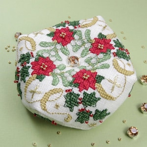 Può includere: Un cuscino per spilli in tessuto bianco con un design natalizio con stelle di Natale, agrifoglio e dettagli dorati. Il cuscino per spilli ha un bottone di cristallo al centro.