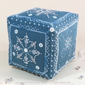 Puede incluir: Cubo de tela azul con diseños de punto de cruz blanco y copos de nieve con cuentas. El cubo tiene detalles de cuentas blancas y lentejuelas. Botones blancos en las esquinas. El tema general es la decoración invernal o festiva.