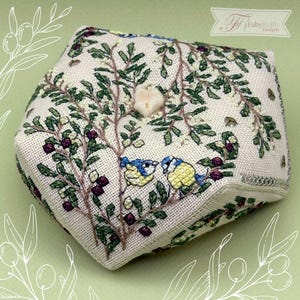 May include: A decorative, hexagonal fabric box with embroidered details. The box features a cream-colored background with green leaves, purple berries, and two blue and yellow birds. A small, white, heart-shaped embellishment is centered on the top.