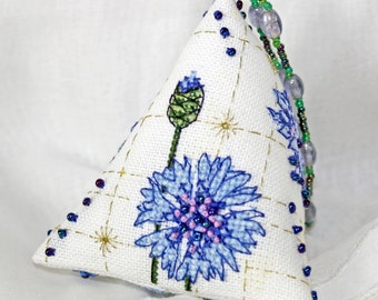 Op de afbeelding: Een wit driehoekig kussen met een geborduurd blauw en groen bloemenontwerp. Het kussen heeft een kralenbies langs de randen.