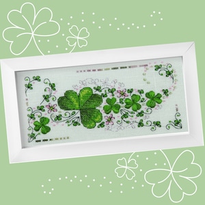 以下が含まれることがあります： 白い額縁に入ったクロスステッチ刺繍。緑の四つ葉のクローバーとピンクの花の模様が施されています。