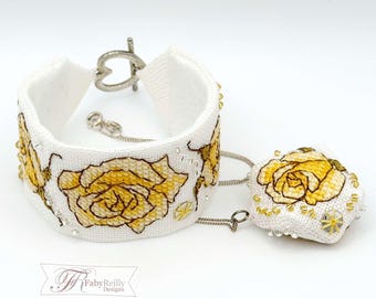 May include: A white fabric cuff bracelet and a matching pendant featuring embroidered yellow roses with brown outlines. The bracelet has a silver-toned heart-shaped toggle clasp, and both pieces are accented with small clear beads and silver chains.