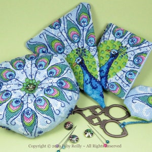 Könnte beinhalten: Ein Set aus vier Nadelkissen aus Nadelmalerei mit einem Pfauendesign in Blautönen, Grün- und Rosatönen. Die Nadelkissen sind herzförmig, quadratisch und zwei dreieckig. Die Nadelkissen sind von Nähzubehör umgeben, darunter eine Schere und Nadeln.