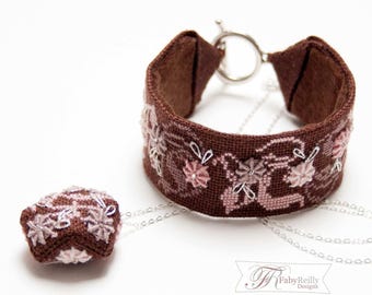 May include: A brown fabric bracelet with a silver toggle clasp and a matching small square pendant on a silver chain. The bracelet and pendant feature intricate cross-stitch embroidery in shades of pink and white, with small silver accents.