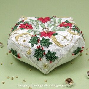 Op de afbeelding: Een wit stoffen naaldkussen met een kerstthema. Het is versierd met rode en groene hulst, witte bloemen en gouden accenten. Het naaldkussen heeft de vorm van een vierkant met afgeronde hoeken.