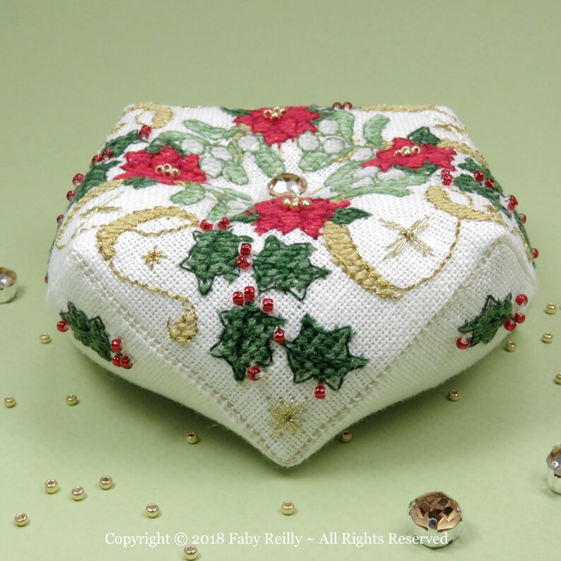 Op de afbeelding: Een wit stoffen naaldkussen met een kerstthema. Het is versierd met rode en groene hulst, witte bloemen en gouden accenten. Het naaldkussen heeft de vorm van een vierkant met afgeronde hoeken.