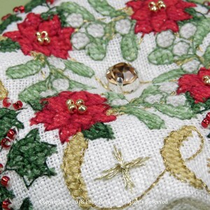 Op de afbeelding: Close-up van een witte stoffen kerstversiering met rode kerststerren, groene hulstbladeren, gouden versieringen en een heldere strasssteen.