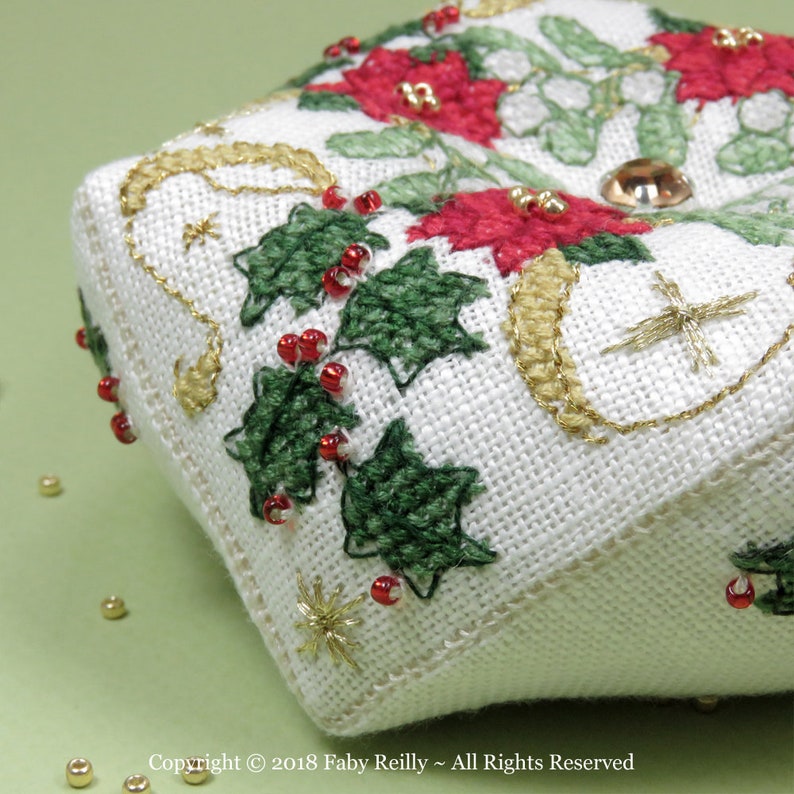 Op de afbeelding: Een witte stoffen kerstversiering met groene hulstbladeren, rode bessen en gouden borduurwerk. De versiering is versierd met kleine gouden kralen en &eacute;&eacute;n grote gouden kraal.