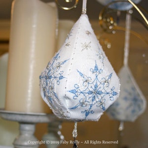 以下が含まれることがあります： 青い雪の結晶の刺繍が施された白い布のクリスマスオーナメント。オーナメントは金色のスタンドから吊り下げられています。