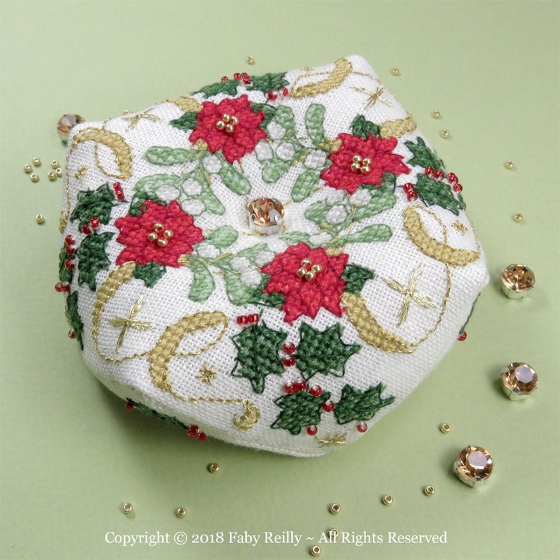 Op de afbeelding: Een wit stoffen naaldenkussen met een kerstontwerp met kerststerren, hulst en gouden accenten. Het naaldenkussen heeft een kristallen knoop in het midden.