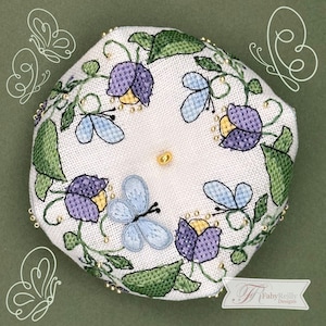 May include: An octagonal white pincushion with floral and butterfly embroidery. The design includes purple and yellow flowers, light blue butterflies, and green leaves, with gold bead accents. The pincushion has a gold centre and is set against a green background.