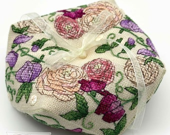 May include: A square, beige fabric pincushion featuring intricate embroidery of pink, peach, and purple flowers with green leaves. A sheer, translucent white ribbon is tied in a bow across the top center. The text Faby Reilly Designs is visible.