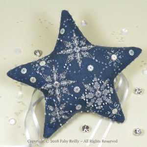 May include: A blue star-shaped pillow with white snowflake embroidery and iridescent sequins. The pillow is decorated with white beads and has a white ribbon attached.