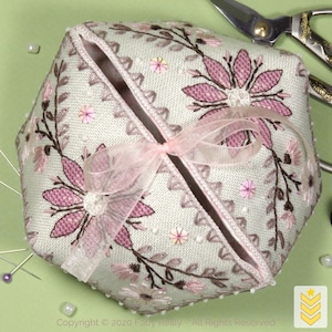 Op de afbeelding: Een lichtgroene, geometrisch gevormde naaldenkoker met roze bloemenborduurwerk en een roze striksluiting. De koker heeft een gedetailleerd ontwerp van bloemen en bladeren, met kleine kralen langs de randen. Schaar en spelden zijn zichtbaar op de achtergrond.