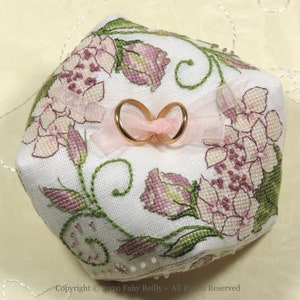 Puede incluir: Un cojín de anillos de tela blanca con flores bordadas en rosa y verde y dos alianzas doradas en la parte superior. El cojín está decorado con pequeñas cuentas blancas alrededor de los bordes. Copyright © 2020 Faby Reilly - Todos los derechos reservados.
