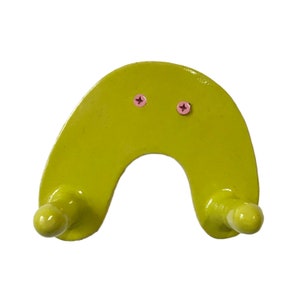 May include: A lime green metal wall hook shaped like a horseshoe with two rounded hooks on the ends. The hook has two pink screws in the center.