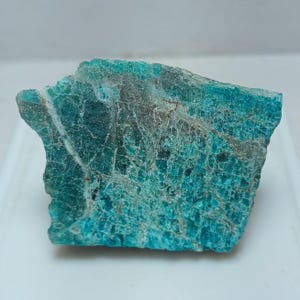 May include: A rough-cut, teal-colored gemstone with a textured surface and visible veining. The stone has a mix of light and dark teal hues, with areas of gray and white. The overall shape is irregular, with a natural, raw appearance.