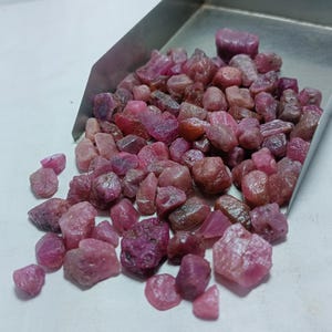 Puede incluir: Primer plano de un montón de piedras preciosas de rubí rosa. Las piedras preciosas son ásperas y sin pulir, y están esparcidas en una bandeja de metal.