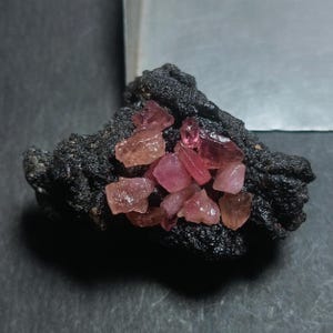 May include: A cluster of rough, pink gemstones rests on a dark, textured rock. The gemstones vary in shade from light pink to deep rose, with a few showing hints of orange. The rock has a porous, almost volcanic appearance.