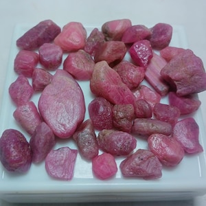 May include: A collection of pink ruby rough gemstones in various sizes and shapes. The rubies are displayed on a white surface.