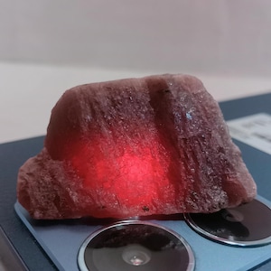 Puede incluir: Un gran cristal de forma irregular, rosa y morado, con un brillo rojo que emana del centro. El cristal está sentado sobre una superficie azul oscuro con dos círculos negros.