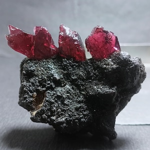 Op de afbeelding: Een mineraalexemplaar met drie doorschijnende, dieprode, grillige kristalformaties die uitsteken bovenop een donkere, poreuze en getextureerde vulkanische rotsbasis met een ruw en onregelmatig oppervlak.