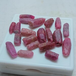 May include: A collection of raw, uncut ruby gemstones in varying shades of pink and red. The gemstones are displayed on a white surface, showcasing their natural, irregular shapes and textures. These rough rubies are ideal for jewelry making or collecting.