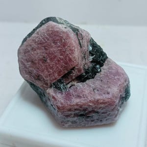 May include: A natural ruby gemstone with a deep red colour, partially encased in black matrix. The rough stone displays a crystalline structure. The stone is set against a white background.