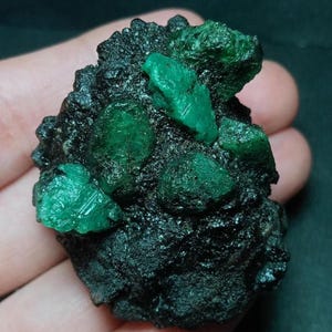 Könnte beinhalten: Ein rohes Mineralexemplar mit mehreren leuchtend grünen Smaragdkristallen, die in eine dunkle, texturierte und poröse schwarze Matrix eingebettet sind. Der Stein zeigt einen starken Kontrast zwischen den hellgrünen Kristallstrukturen und dem rauen Gestein.