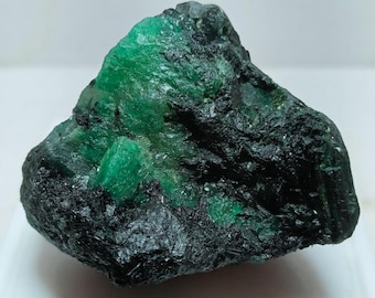 May include: A raw mineral specimen featuring a combination of deep, dark black rock matrix and vibrant, translucent emerald green crystal formations. The surface displays a rough, jagged texture with contrasting color zones.