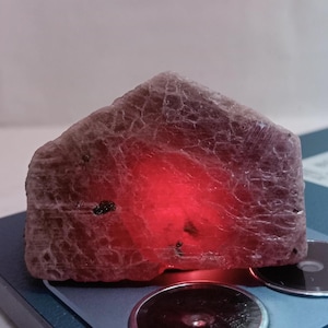 Peut inclure: Une grande pierre précieuse brute, rose et rouge, avec un centre rouge translucide, presque incandescent. La pierre précieuse est posée sur une surface bleu foncé.