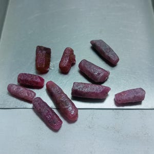 May include: A collection of ten rough, pink ruby gemstones. The gemstones are various sizes and shapes, with some being long and thin, while others are shorter and wider.