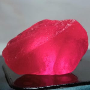 May include: A raw, translucent gemstone with a vibrant, saturated hot pink color. The stone features an irregular, organic shape with multifaceted, textured surfaces that catch the light, creating varying depths of pink and bright red tones.