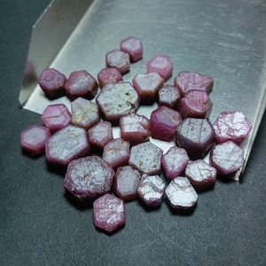 Könnte beinhalten: Eine Sammlung von rosafarbenen Rubingranaten, jeder mit einer sechseckigen Form und einer facettierten Oberfläche. Die Rubine sind auf einem silbernen Tablett zu einem Haufen angeordnet.