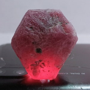 Op de afbeelding: Een grote, gefacetteerde, roze robijn edelsteen met een ruwe, natuurlijke afwerking. De robijn wordt van onderen belicht, waardoor zijn dieprode kleur wordt benadrukt.