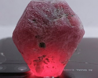 May include: A large, faceted, pink ruby gemstone with a rough, natural finish. The ruby is illuminated from below, highlighting its deep red color.