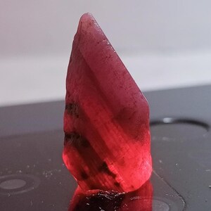 May include: A raw, uncut ruby gemstone. The ruby is a deep red color with a slightly translucent appearance. The stone has a rough, textured surface and a pointed top. The gemstone is displayed against a dark background.