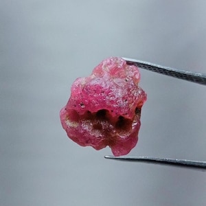 May include: A rough, pink ruby gemstone held by tweezers. The ruby has a deep, rich pink color and an irregular shape.