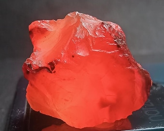 Könnte beinhalten: Ein roher, ungeschliffener Edelstein mit einem leuchtenden orange-roten Farbton. Die raue Oberfläche des Steins ist beleuchtet und zeigt seine natürliche Textur und kristalline Struktur. Der Edelstein ist wahrscheinlich ein Mineral, möglicherweise für Schmuck oder Sammlungen.